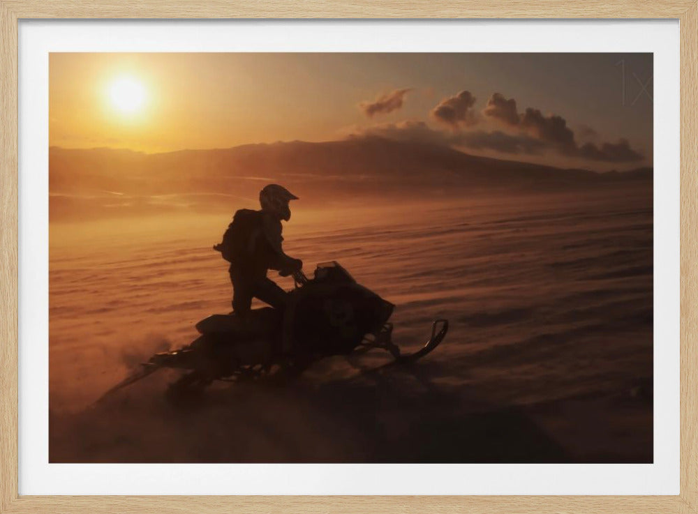 A silhouette of a person wearing a helmet and backpack stands on a snowmobile in a vast, snowy landscape during a golden sunset. The wind blows snow across the ground, creating a hazy, atmospheric effect, with mountains visible in the distance under a warm sky. Wall Art
