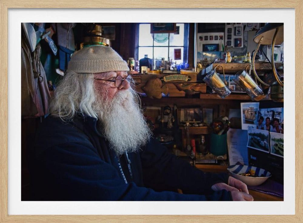 A profile portrait of an elderly man with a long, full white beard and hair, wearing glasses and a cream-colored knit beanie. He sits indoors in a warmly lit, cluttered room that looks like a cabin or a pub, with his hands clasped in front of him, gazing thoughtfully to the side. Poster