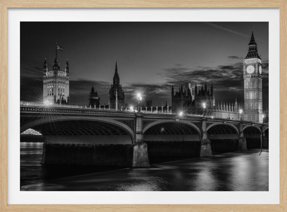 A framed, black and white nighttime photograph of Westminster Bridge in London. The iconic Houses of Parliament and the glowing Big Ben clock tower stand prominently in the background against a dark, cloudy sky. The streetlights on the bridge create starburst effects, and their reflections shimmer on the surface of the River Thames in the foreground. Poster