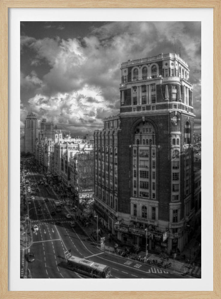 A dramatic black and white high-angle photograph of a bustling city street. A large, ornate historical building dominates the right side, while traffic flows along the avenue on the left under a sky filled with heavy, textured clouds. Poster