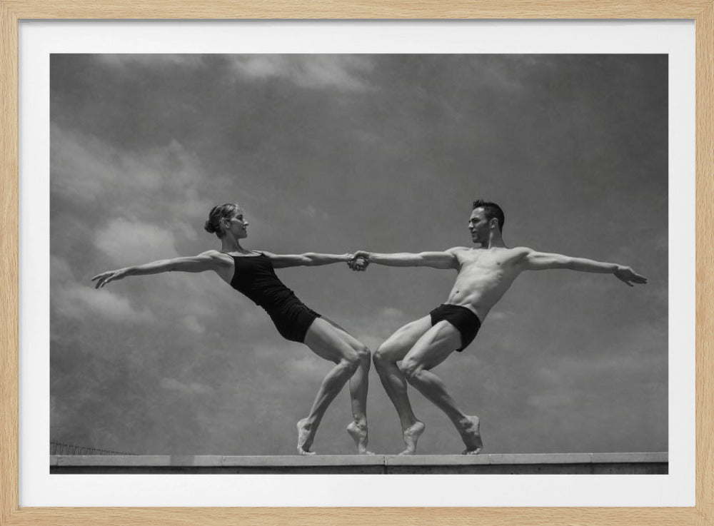 A black and white photograph of a male and female ballet dancer striking a symmetrical, athletic pose on a ledge. They hold hands, leaning away from each other in a display of strength and balance against a cloudy sky, all enclosed within a silver frame. Print