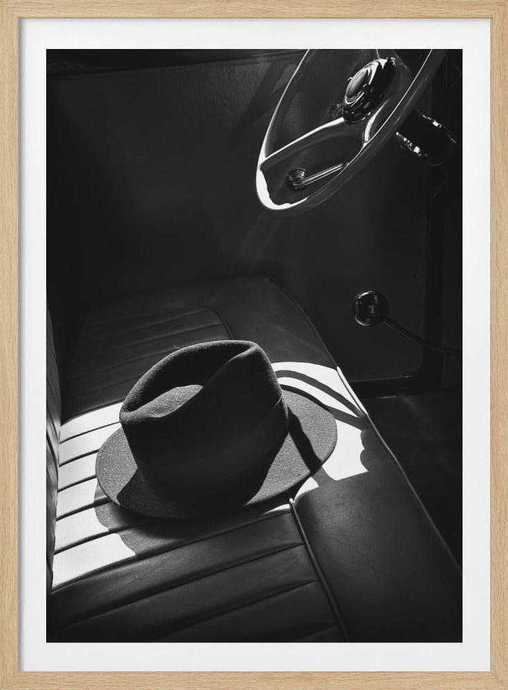 A black and white photograph of a dark fedora hat resting on the passenger seat of a vintage car. Sunlight streams through the window, casting strong shadows and highlighting the ribbed texture of the seat and parts of the steering wheel in the background. Print