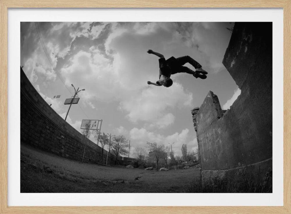 A dynamic black and white fisheye photograph of a person mid-flip, jumping over a crumbling wall in an urban setting under a cloudy sky. The image has a silver-colored frame. Print