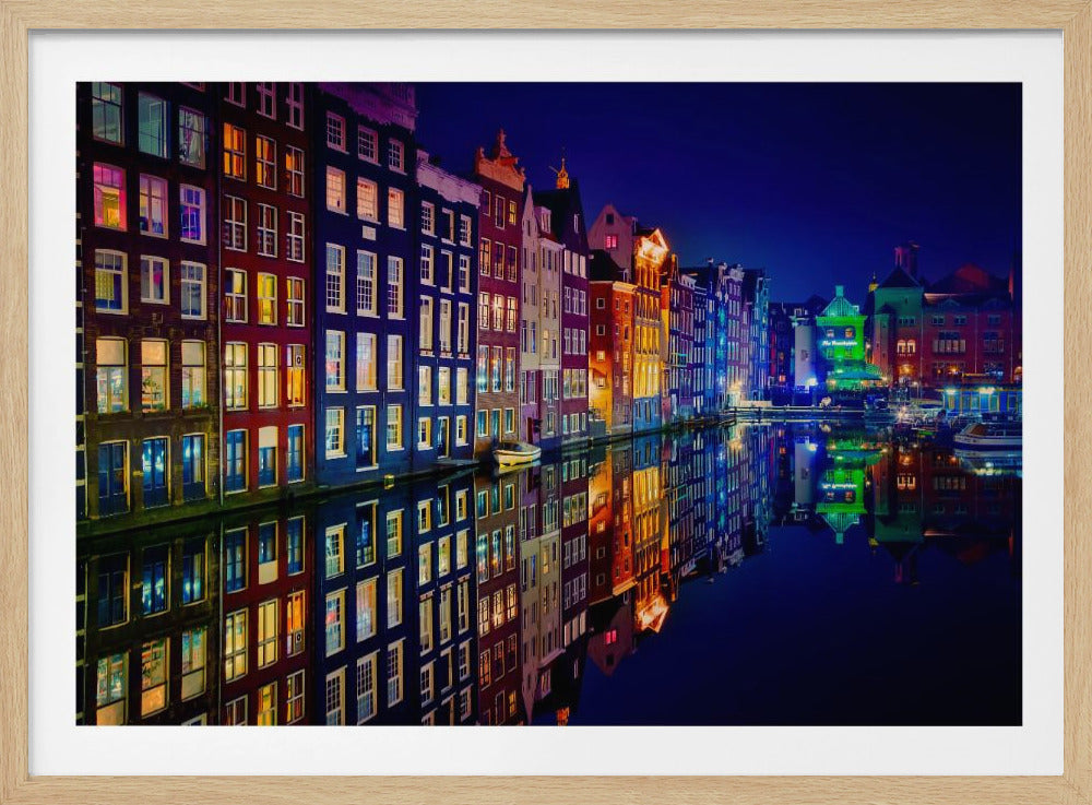 A vibrant nighttime photograph of colorful Dutch canal houses in Amsterdam, with their illuminated windows casting a perfect, colorful reflection on the still water of the canal below. The scene is captured under a dark blue night sky and is presented within a silver frame. Poster