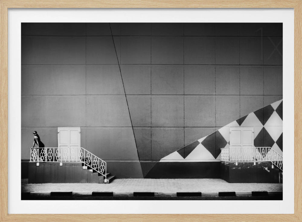 A framed black and white photograph of a modern urban building. A woman in a dark outfit stands on the left by a white ornate railing and door. The large gray wall behind her is bisected by strong diagonal lines and features a bold black and white diamond pattern on the right, where another door and railing are visible. Decor