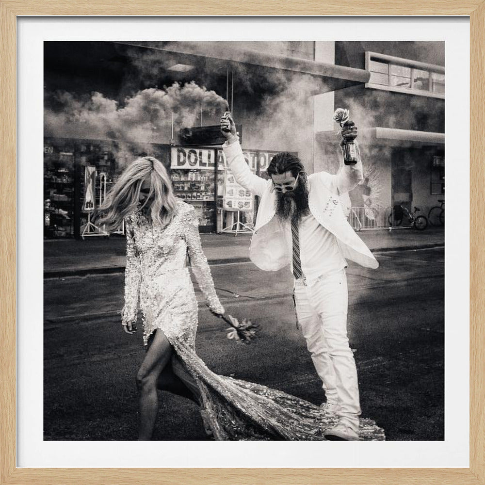 A black and white photograph in a light wood frame showing a stylish couple walking down a street. The woman wears a sparkling sequin gown, and the man, in a white suit with a long beard, triumphantly raises his arms, holding a smoke bomb and a bottle with a rose. Decor