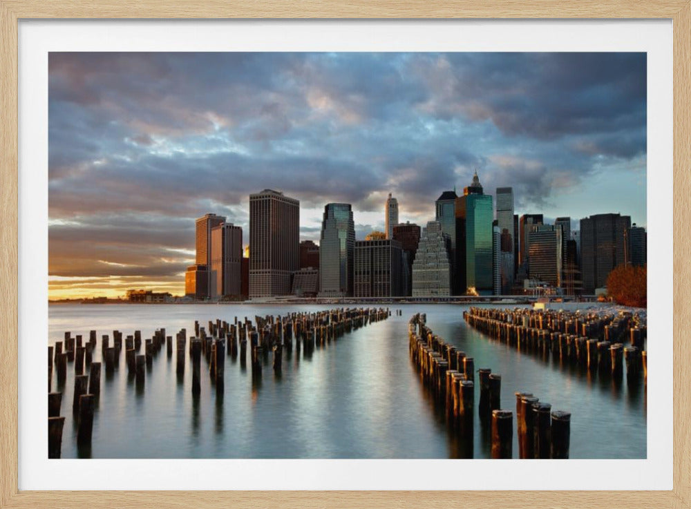 A framed photograph of the Lower Manhattan skyline at sunset, viewed from across the water. Rows of old wooden pier pilings lead the eye towards the city's skyscrapers, which are illuminated by the warm light of the setting sun under a dramatic, cloudy sky. Decor