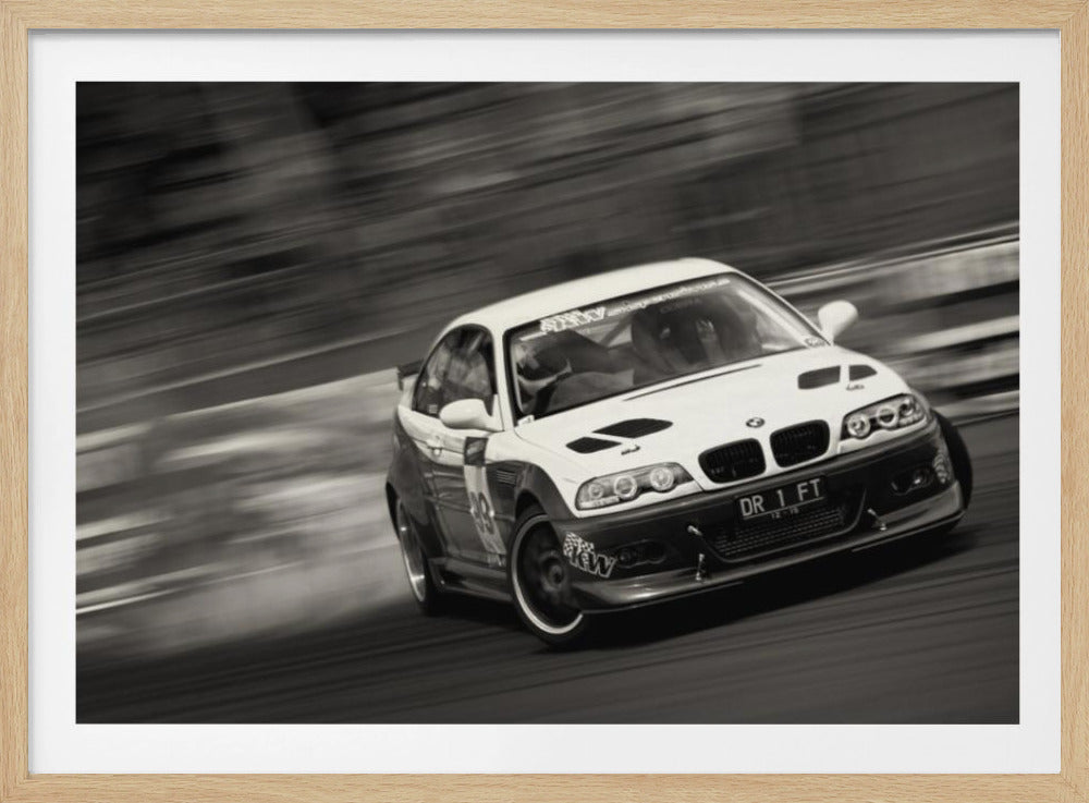 A framed, black and white action photograph of a white and black BMW race car drifting at high speed around a corner. The background is blurred with motion lines, emphasizing the car's speed as it turns, and a puff of smoke is visible from the tires. Wall Art