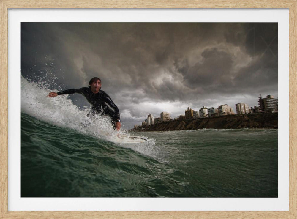 A close-up, low-angle shot of a male surfer in a black wetsuit riding a dark green ocean wave. In the background, a city skyline sits atop a cliff under a dramatic, dark, and stormy sky. The image is presented within a silver picture frame. Print