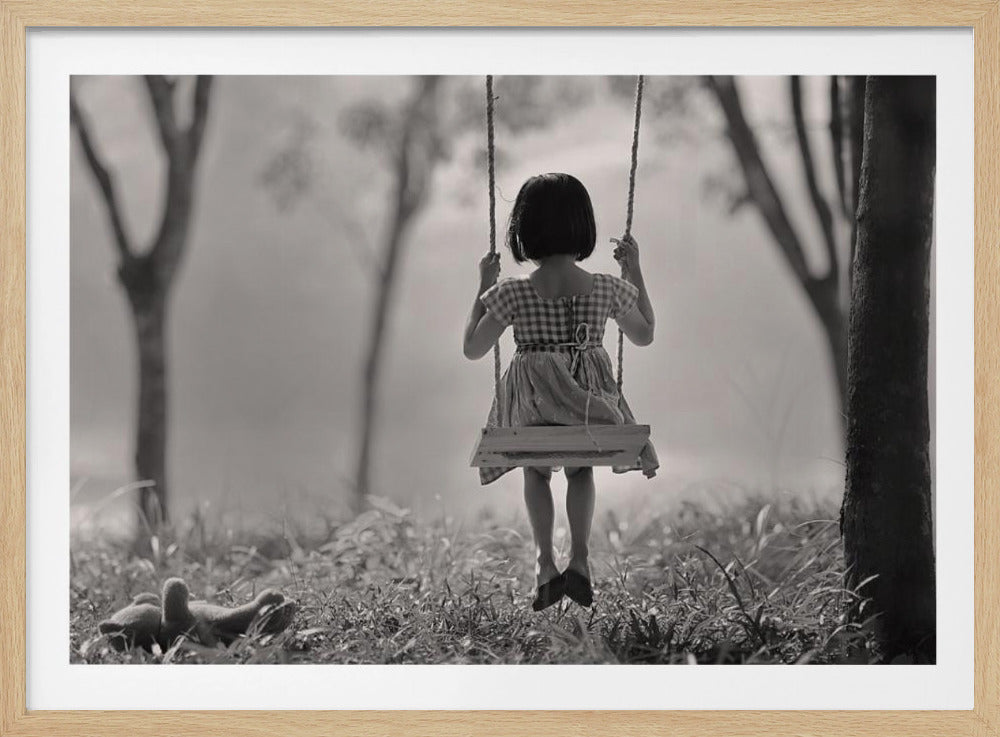 A black and white photograph of a young girl from behind, swinging on a rope swing in a grassy field. She has short dark hair and wears a patterned dress. Small goslings are visible in the grass to the left, and trees are blurred in the background, creating a nostalgic and peaceful scene. The image is presented in a silver frame. Artwork