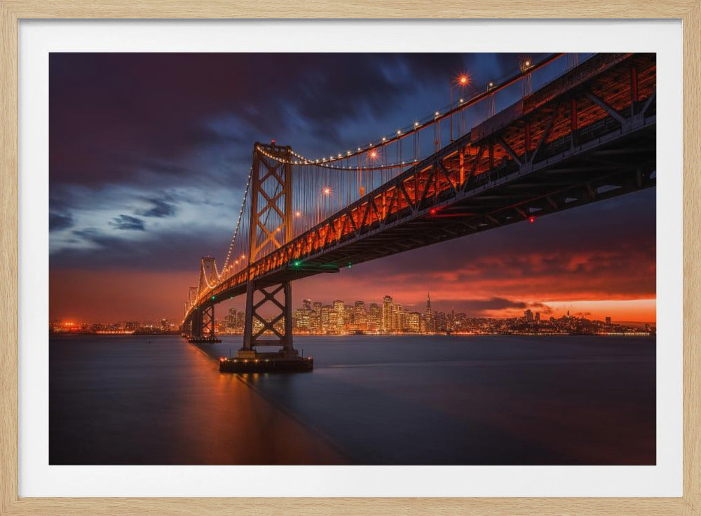 A stunning low-angle, long-exposure photograph of the illuminated San Francisco-Oakland Bay Bridge at dusk, with the city skyline lit up in the background under a dramatic red and dark blue sunset sky. The calm water of the bay reflects the warm lights of the bridge and the cityscape. Artwork
