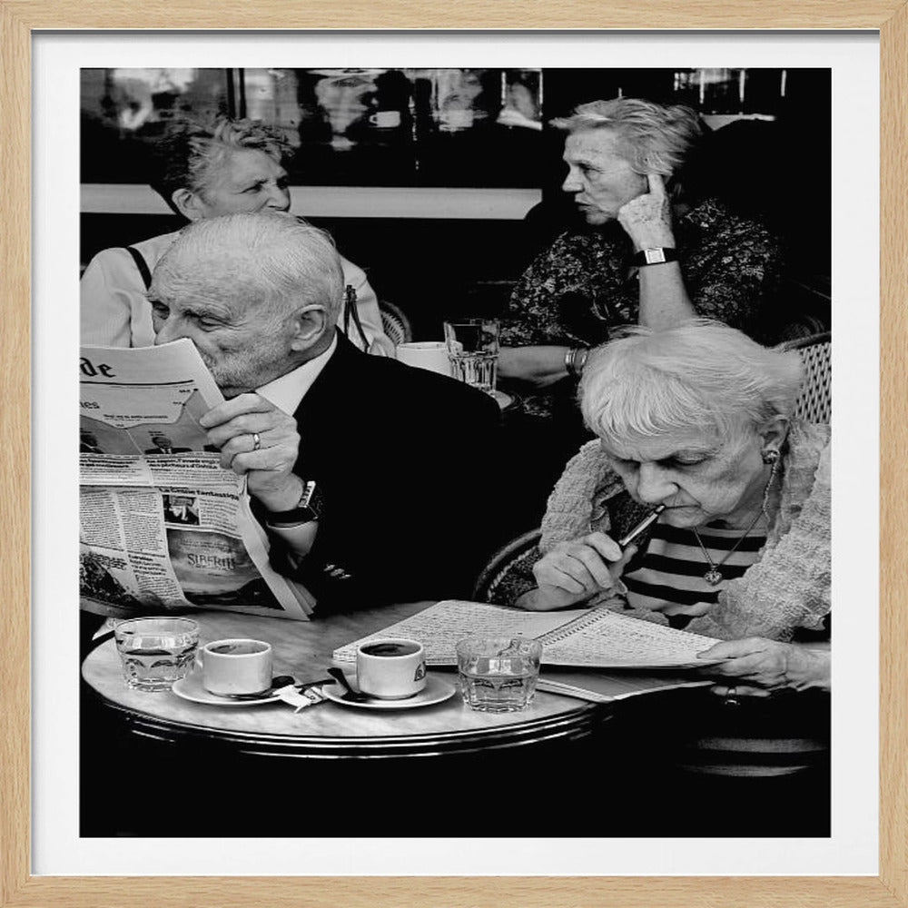 A black and white photograph of four elderly people sitting at a table in a cafe. A man in the foreground is reading a 'Le Monde' newspaper, while a woman beside him is focused on writing in a notebook. Two other women are seated in the background, engaged in conversation. Wall Art