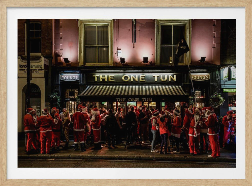 A nighttime photograph of a large crowd of people dressed in Santa Claus costumes standing outside a warmly lit pub called 'The One Tun', creating a festive holiday atmosphere on a city street. Print