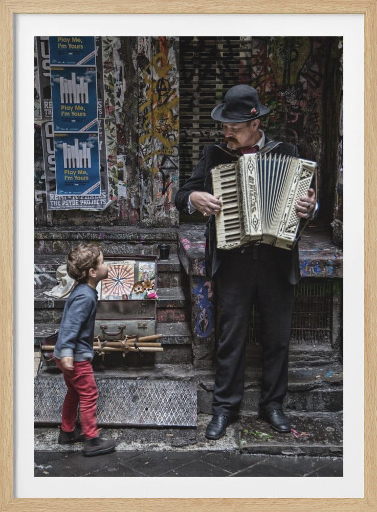 A street musician wearing a black suit and fedora plays a white accordion while standing in a gritty, graffiti-covered alleyway. A small child in red pants and a blue long-sleeved shirt stands on the steps, looking up at the musician with wonder. Decor