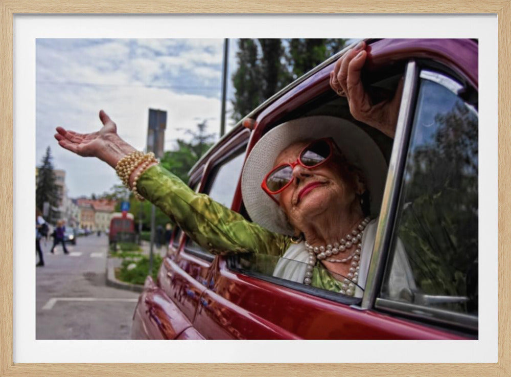 A stylish elderly woman wearing a white hat, red sunglasses, and pearl necklaces leans out of the passenger window of a classic red car. Her arm is outstretched with a joyful expression on her face, enjoying a ride through a city street. Poster