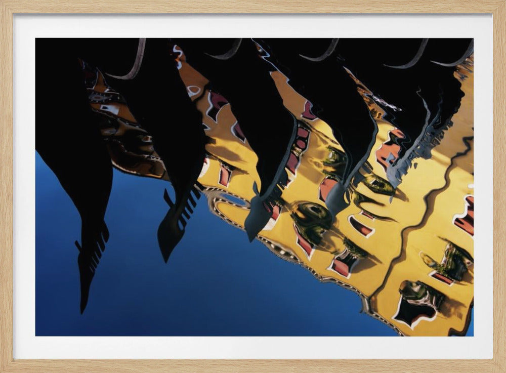 An abstract photograph of a distorted reflection in water, showing a vibrant yellow building against a deep blue sky, with the dark silhouettes of several Venetian gondola prows cutting across the top of the image. The entire scene is enclosed in a silver picture frame. Print