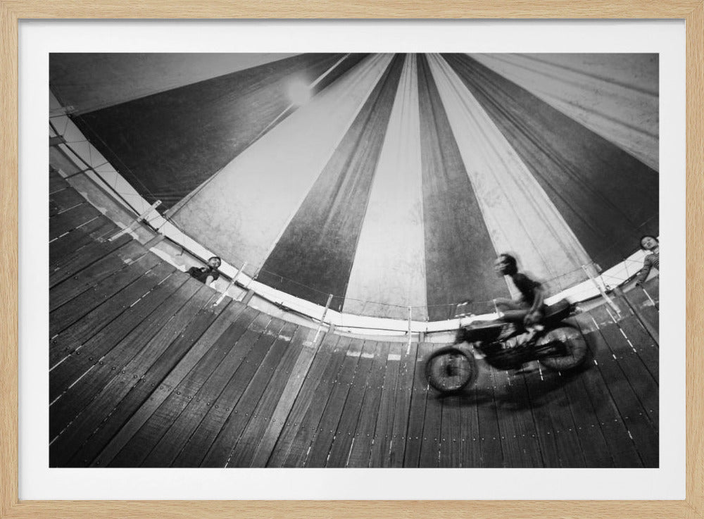 A dynamic, wide-angle black and white photograph of a motorcycle rider performing a stunt in a motordrome, commonly known as the 'wall of death'. The rider is a blur of motion as they speed along the steep, curved wooden walls. The perspective looks up towards the striped canvas top of the tent, with a few spectators visible watching from the railing at the top edge. The entire image is enclosed in a silver frame. Print