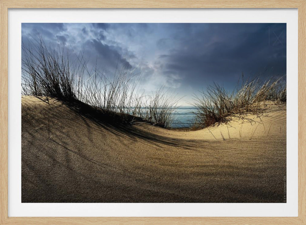 A landscape photograph of a sand dune in the foreground with long grasses casting shadows, looking out to the ocean under a dark and stormy sky. The image is presented within a silver frame. Decor