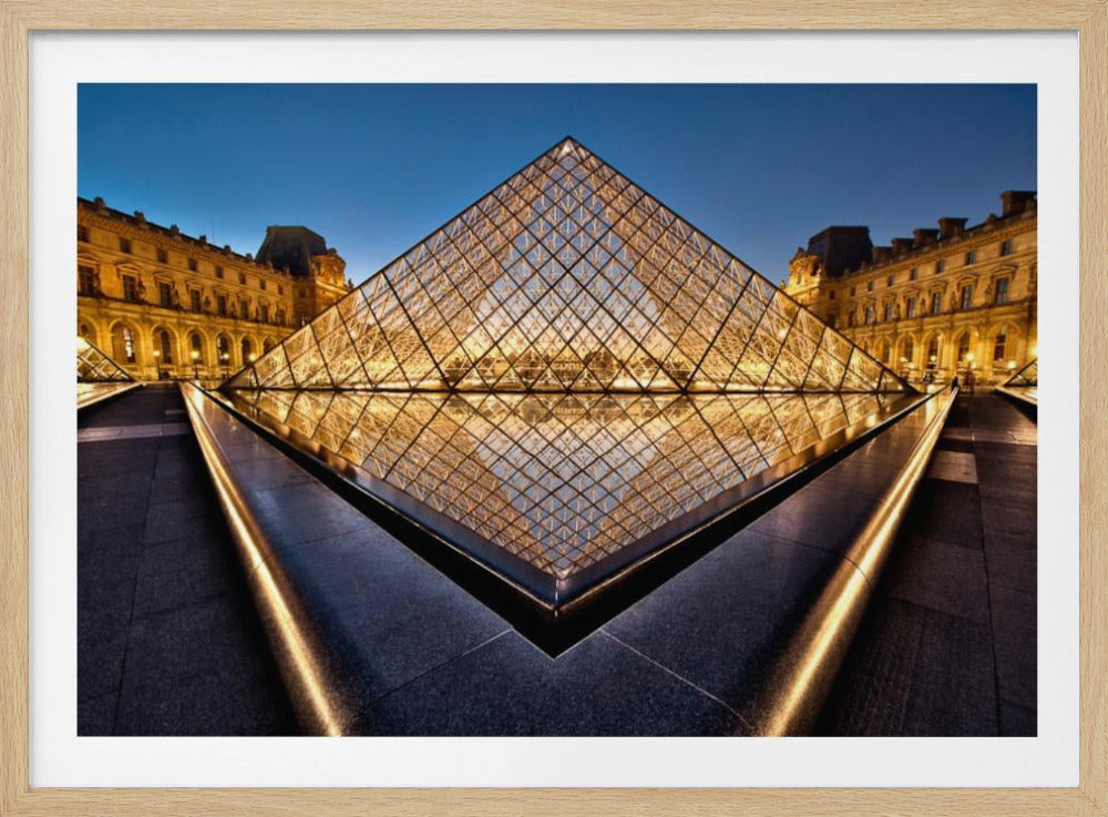 A low-angle, symmetrical photograph of the illuminated Louvre Pyramid at dusk, its golden light reflecting in a dark, still pool of water in the foreground. The historic Louvre Museum buildings are visible in the background against a deep blue evening sky. The entire image is presented within a silver frame. Print