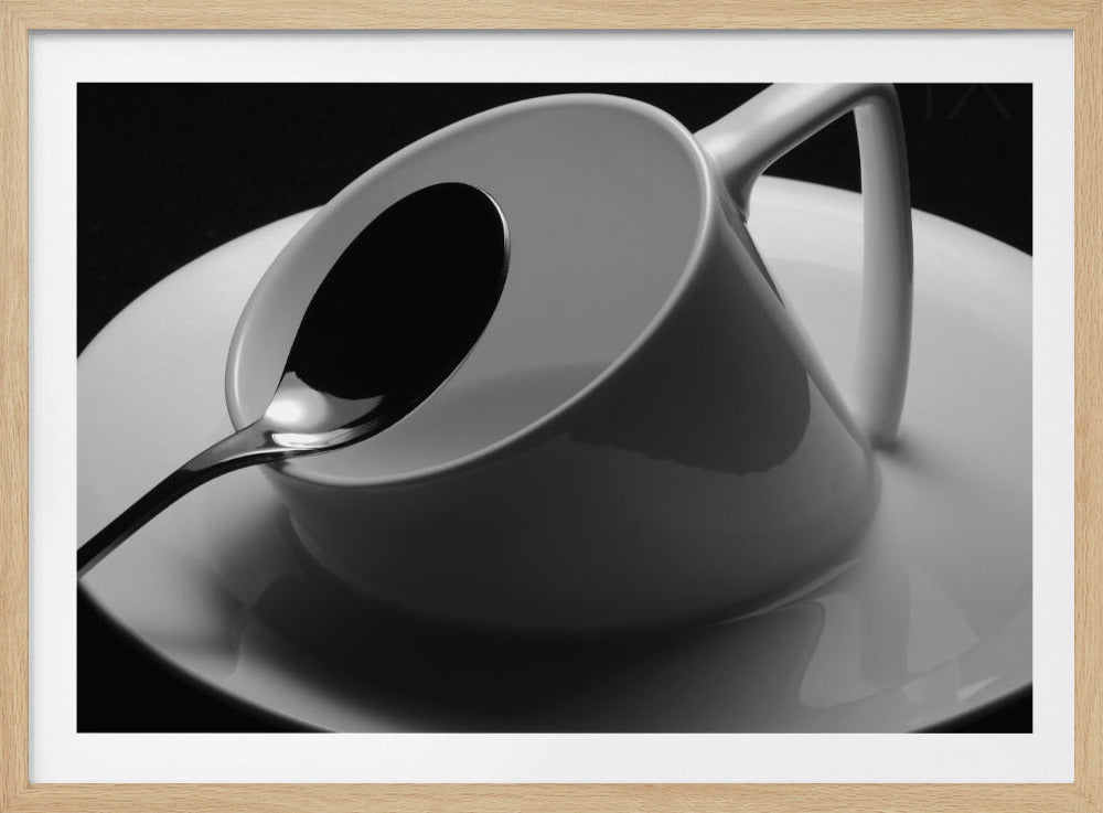 A dramatic black and white close-up photograph of a white porcelain coffee cup tilted on its saucer, with a reflective spoon resting inside. The image has high contrast against a dark background and is presented within a silver-colored frame. Decor