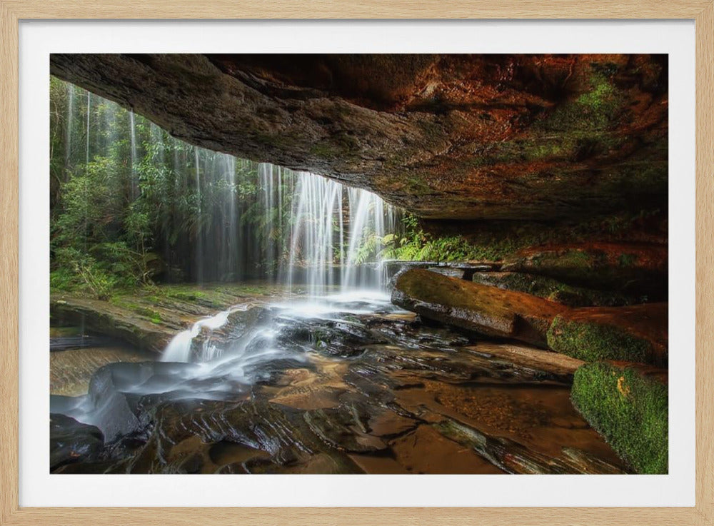 A long-exposure photograph taken from underneath a rock overhang, looking out through a cascading waterfall. The water appears silky as it flows over mossy rocks in a stream below. The scene is framed by a silver border. Print