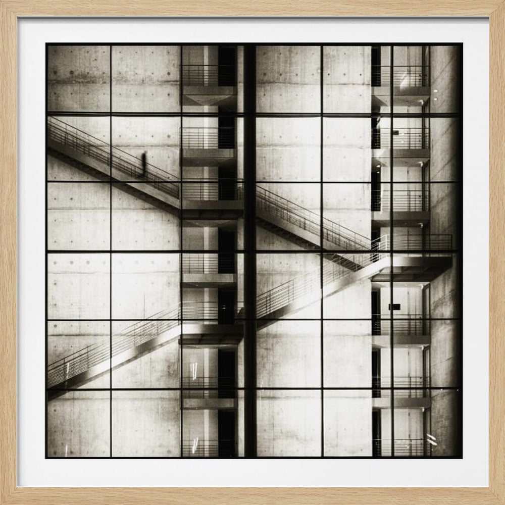 A framed black and white photograph of a modern concrete building's interior, seen through a grid of window panes. The image captures multiple levels of staircases and walkways crisscrossing in a complex, geometric pattern. A shadowy, blurred figure of a person can be seen walking up a staircase on the upper left. Wall Art