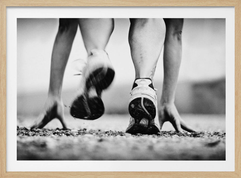A low-angle, black and white action shot of a runner's legs and hands at the starting position on a gravel track. One foot is planted firmly while the other is in motion, blurred as they push off to start a race. Decor