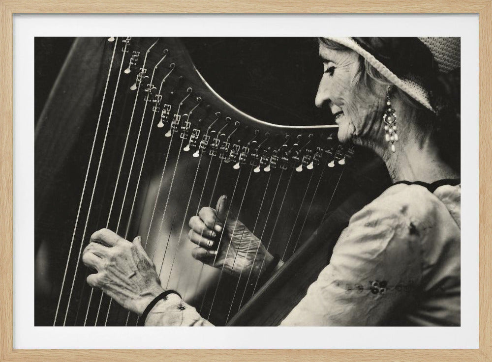 A close-up, black and white photograph of an elderly woman in profile, smiling as she plays a large wooden harp. She wears a light-colored hat and ornate pearl earrings, and her experienced hands are shown plucking the strings. Poster