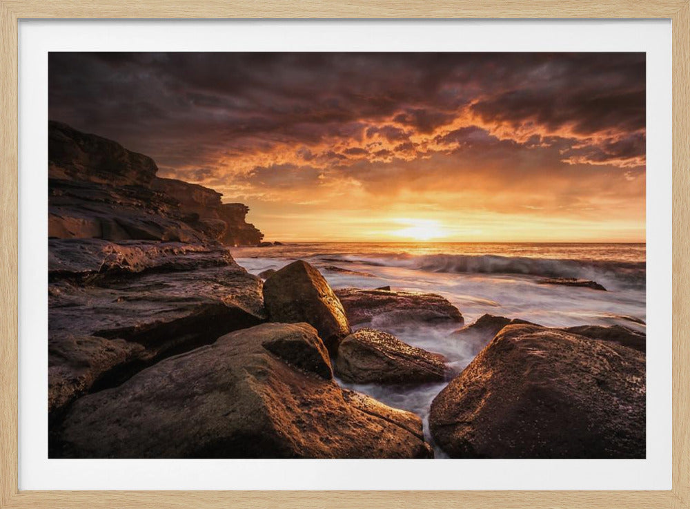 A framed photograph of a dramatic ocean sunset, with fiery orange clouds filling the sky. Large, dark rocks sit on the shore as long-exposure waves create a misty effect around them. Print