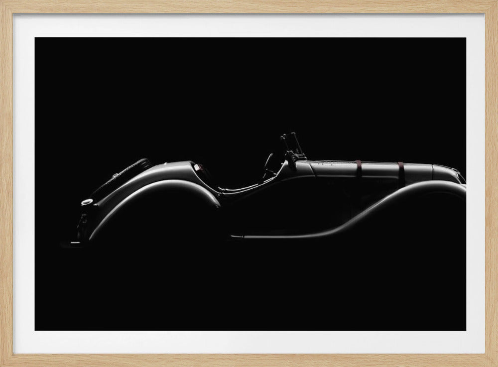 A dramatic black and white photograph showcasing the elegant silhouette of a vintage roadster. The side profile of the classic car is highlighted by subtle light, emphasizing its long hood and sweeping fenders against a solid black background, all within a brushed silver frame. Print