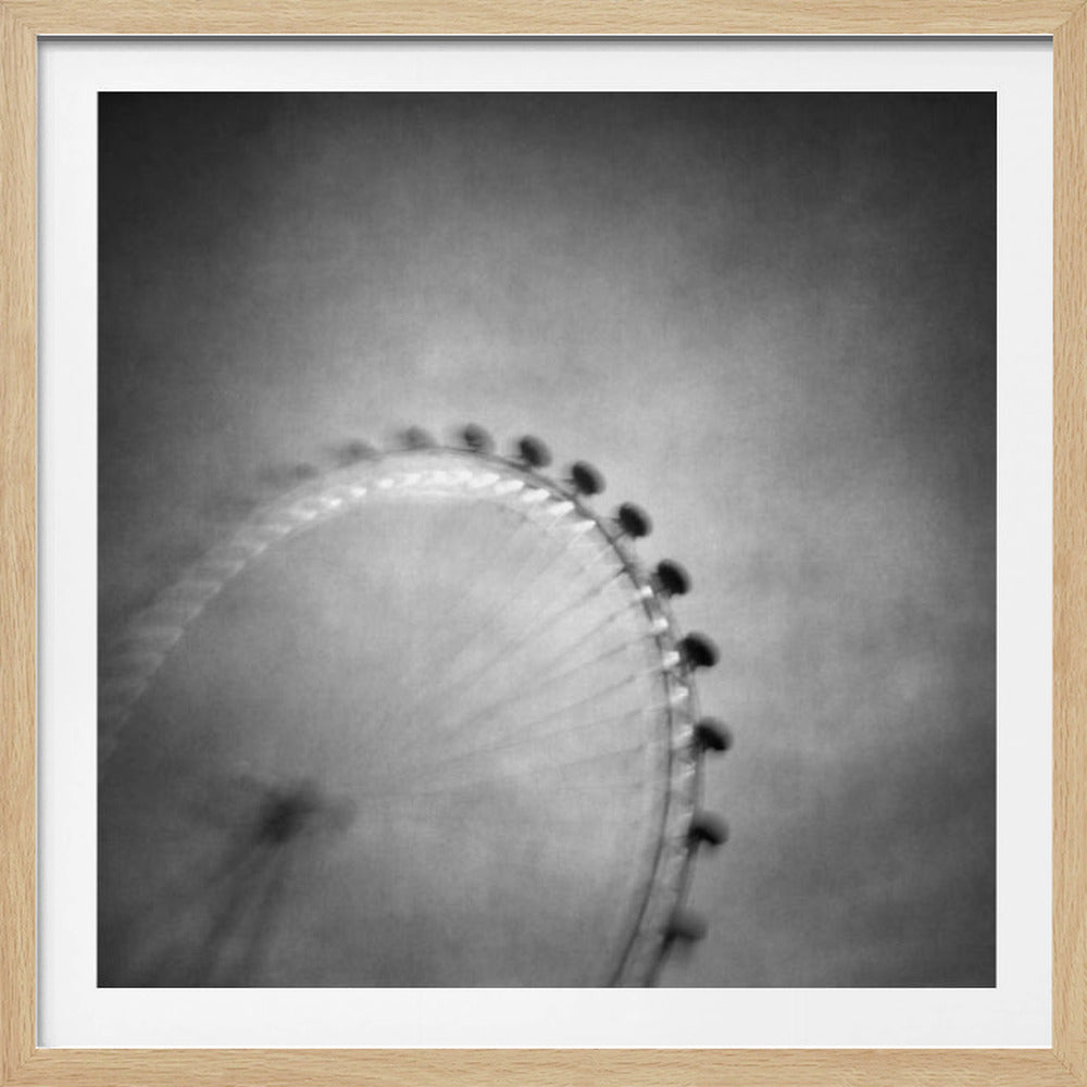 A moody, black and white long-exposure photograph of a large Ferris wheel, creating a soft, blurry effect of its rotation against a grainy gray sky. The artwork is presented in a simple, light wood frame. Poster