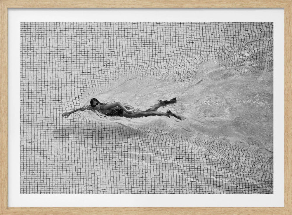 A framed, black and white overhead photograph of a woman swimming freestyle in a tiled pool. She wears dark goggles, and her movement creates a wake that distorts the gridded pattern of the pool floor. Wall Art