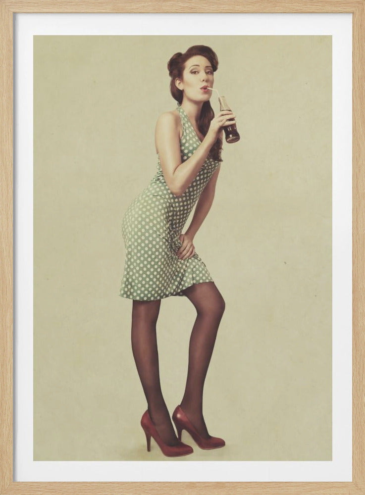 A full-length vintage-style photograph of a pin-up model wearing a green and white polka dot dress, black stockings, and red heels. She poses against a beige background while sipping a dark soda from a glass bottle with a straw. Print