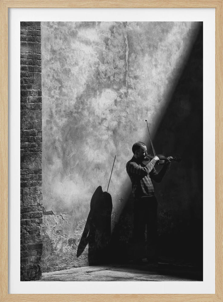 A dramatic black and white photograph of a man playing the violin, standing in a corner against a weathered wall. He is bathed in a sharp diagonal ray of light, casting a long, dark shadow behind him. Artwork