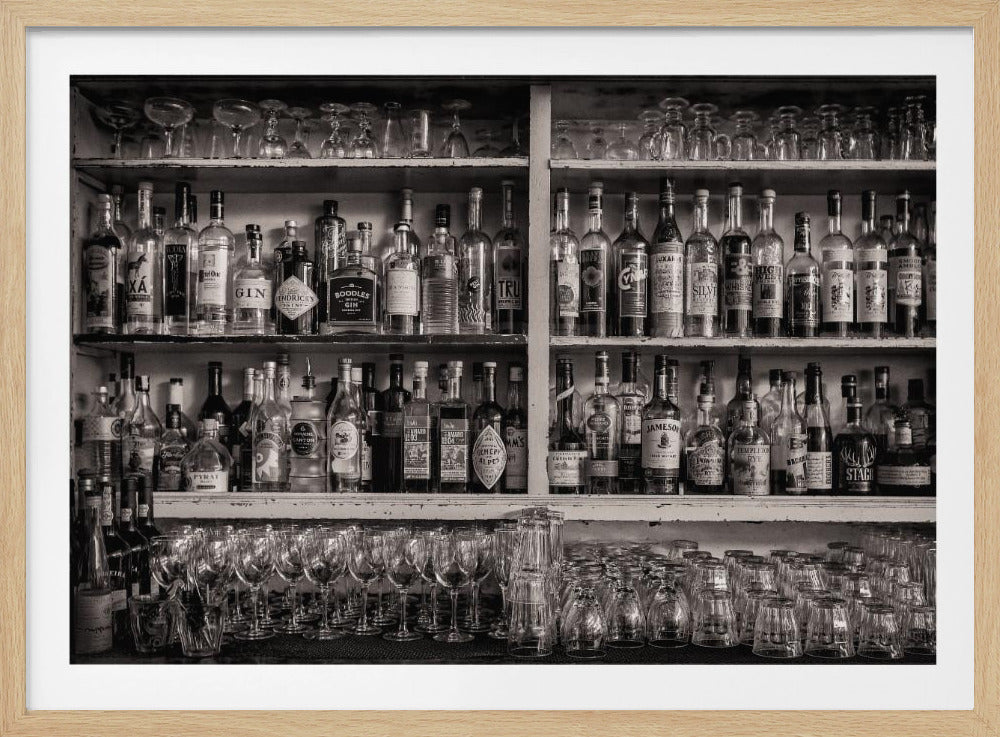 A black and white photograph of a well-stocked bar. Multiple shelves are filled with a wide variety of liquor bottles, and below them, rows of clean, empty wine glasses and tumblers are arranged, all presented within a silver frame. Print