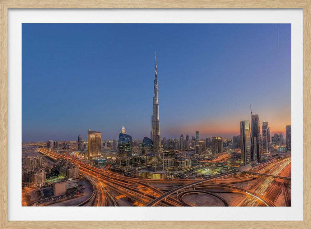 An aerial photograph of the Dubai skyline at dusk, centered on the illuminated Burj Khalifa. The sky transitions from deep blue to warm orange at the horizon, while a complex network of highways below glows with the light trails of traffic. Artwork