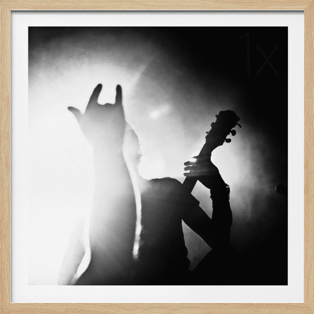 A dramatic black and white photograph of a music concert. A person is silhouetted against a bright stage light, holding up a guitar neck with one hand while another hand is raised in a 'rock on' gesture, capturing the energy of a live performance. Poster