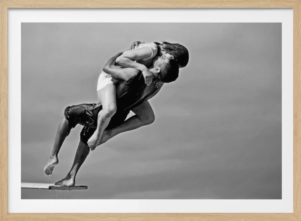 A framed, black and white photograph capturing a dynamic and romantic moment. A man is leaping off a diving board, carrying a woman who is wrapped around him in a passionate embrace, their faces close together. The background is a simple, plain gray, emphasizing their mid-air pose. Wall Art