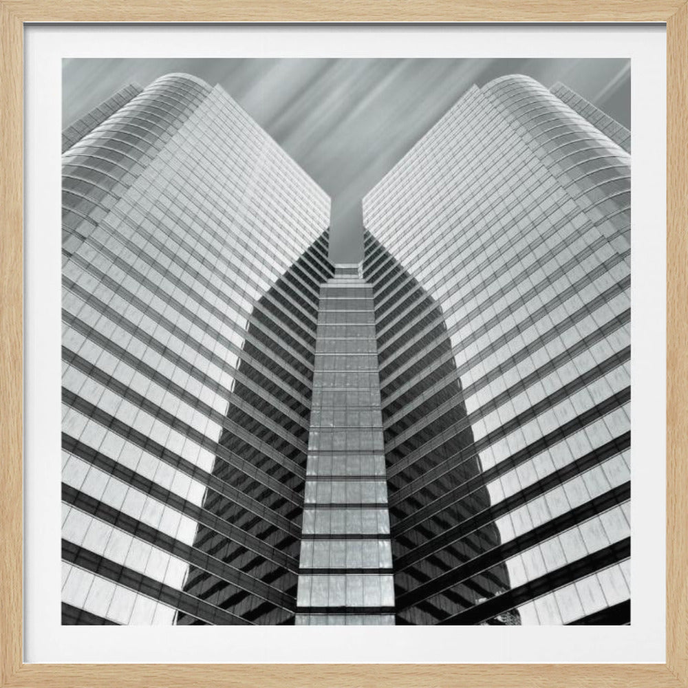 A low-angle, symmetrical black and white photograph of a modern glass skyscraper. The two towers of the building converge towards the center, creating a V-shape against a sky with motion-blurred clouds. The photo is enclosed in a light-colored wooden frame. Print