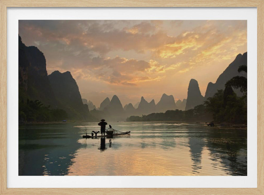 A serene landscape photograph of a silhouetted cormorant fisherman on a bamboo raft, floating on a calm river at sunrise or sunset. In the background, majestic karst mountains rise into a cloudy, golden-orange sky, with the warm light reflecting on the water's surface. Decor