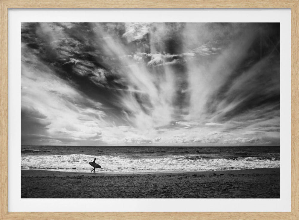 A dramatic black and white photograph of a lone surfer silhouetted against the sea, walking along a sandy beach. The sky is filled with spectacular, radiating clouds that stretch across the horizon, creating a powerful and moody atmosphere. The image is enclosed in a silver frame. Poster