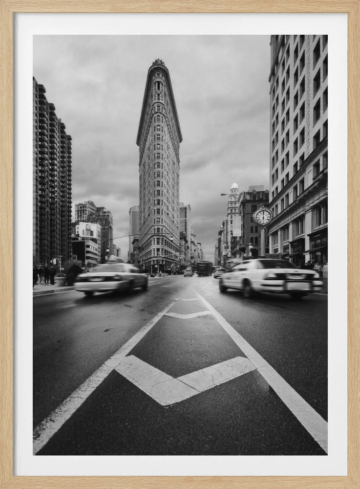 A dynamic black and white, low-angle photograph of the iconic Flatiron Building in New York City. The street in the foreground has bold white lines leading the eye to the building, while cars on either side are blurred with motion, capturing the city's fast pace under a cloudy sky. Decor