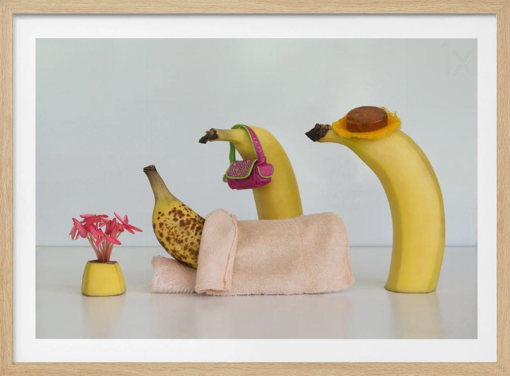 A whimsical still life photo featuring three bananas personified as a family or friends. One overripe, spotted banana lies down, covered by a pink towel, while another banana with a tiny pink purse and a third with a small hat seem to be visiting. A small vase with pink flowers sits nearby on the white surface. Poster