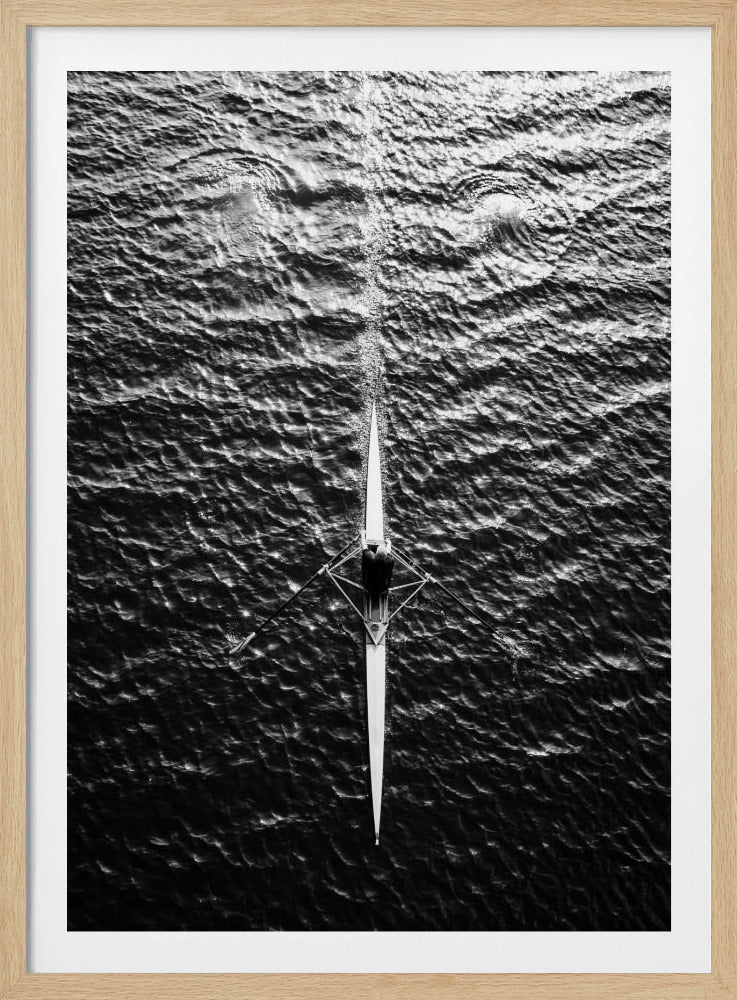 A dramatic black and white overhead photograph of a lone rower in a sleek racing shell, cutting a straight path through dark, textured water. The high-contrast lighting highlights the ripples on the water's surface and the rower's focused form. Artwork