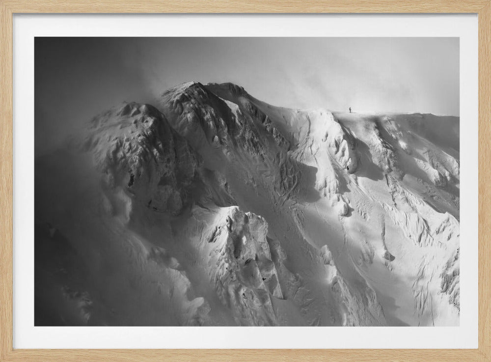 A dramatic black and white photograph of a person standing alone on the ridge of a massive, snow-covered mountain, emphasizing the scale and solitude of the vast, rugged landscape, all presented in a silver frame. Wall Art