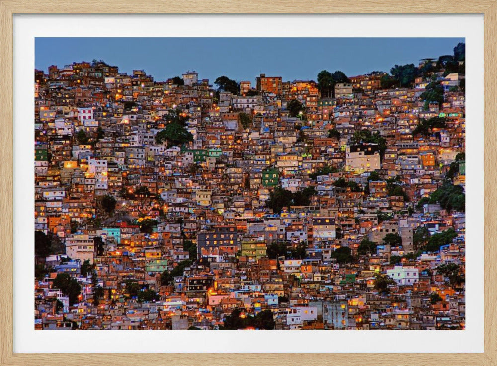 A photograph of a densely packed favela built on a hillside at dusk. The numerous small, colorful houses are illuminated from within, casting a warm orange glow that contrasts with the deep blue of the twilight sky. The image is presented within a silver picture frame. Artwork