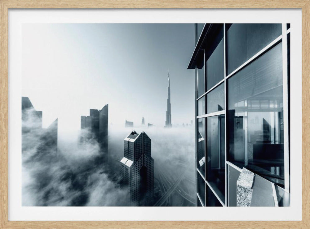 A framed black and white photograph with a cool blue tint, depicting the Dubai skyline from a high-rise window, with skyscrapers including the Burj Khalifa piercing through a thick blanket of fog. Artwork