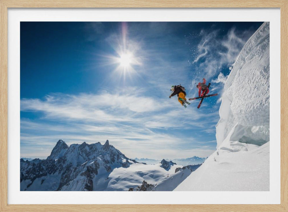 Two skiers in bright gear are captured mid-air as they jump off a steep, snowy cliff under a brilliant sun. The background features a vast expanse of a blue sky with wispy clouds and a majestic snow-covered mountain range. Print