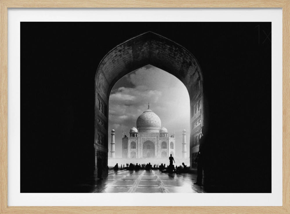 A dramatic black and white photograph of the Taj Mahal seen through a large, dark archway which frames the view. The iconic mausoleum is in the distance under a cloudy sky, and the silhouettes of many people are scattered on the wet, reflective ground in the foreground. Decor
