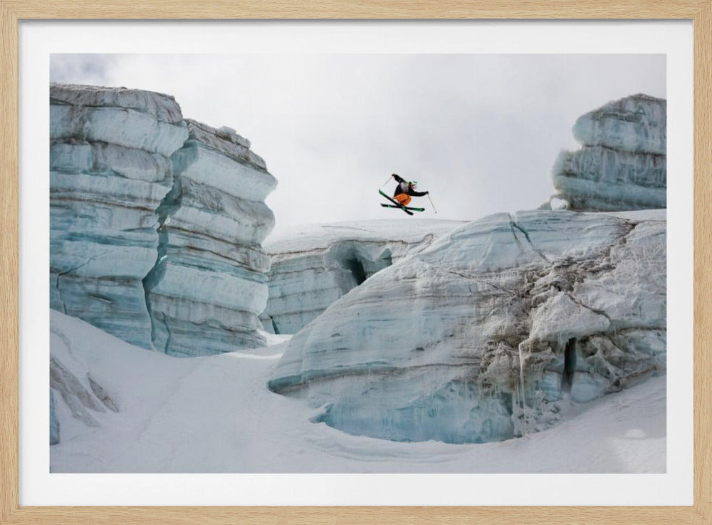 A framed photo of a skier in mid-air, jumping over a snowy crevasse between large, layered glacier formations under an overcast sky. The skier wears a black top and orange pants. Wall Art